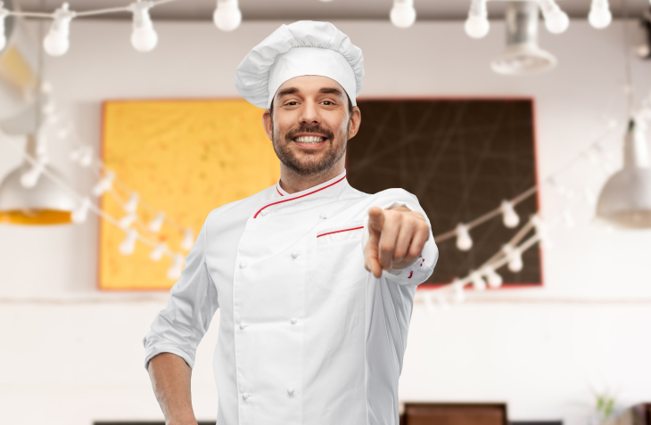 un cuisinier avec sa toque, souriant et pointant du doigt